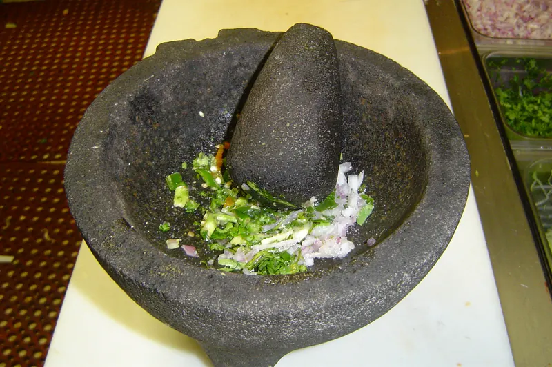 A stone molcajete and pestle with chopped onion, cilantro and jalapeño, guacamole being made