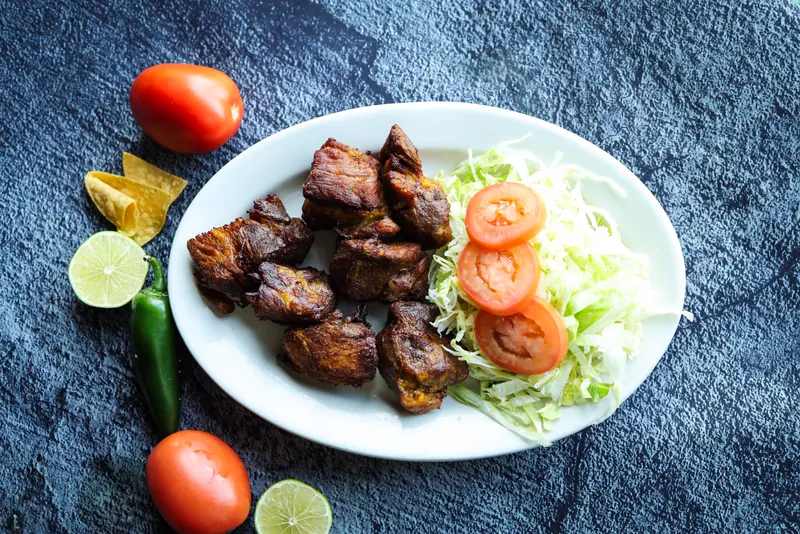 Carnitas de puerco with shredded lettuce and tomato slices on a white oval plate, with limes, a jalapeño and tomatoes on slate