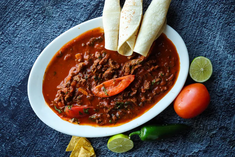 Beef in a red chile sauce with a tomato wedge and three rolled flour tortillas on a white oval plate, limes and a jalapeño beside it