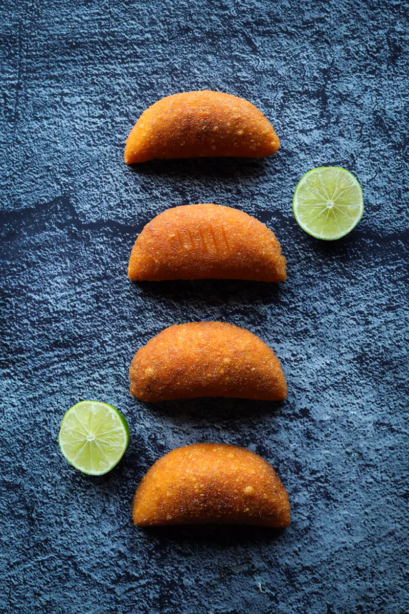Four golden fried empanadas in a row with two halved limes on blue-grey slate