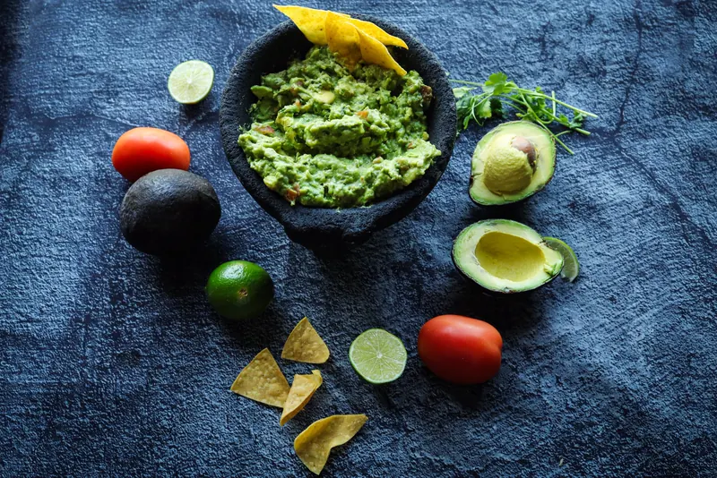 Guacamole in the black molcajete with halved avocados, limes, tomatoes, cilantro and tortilla chips on blue-grey slate