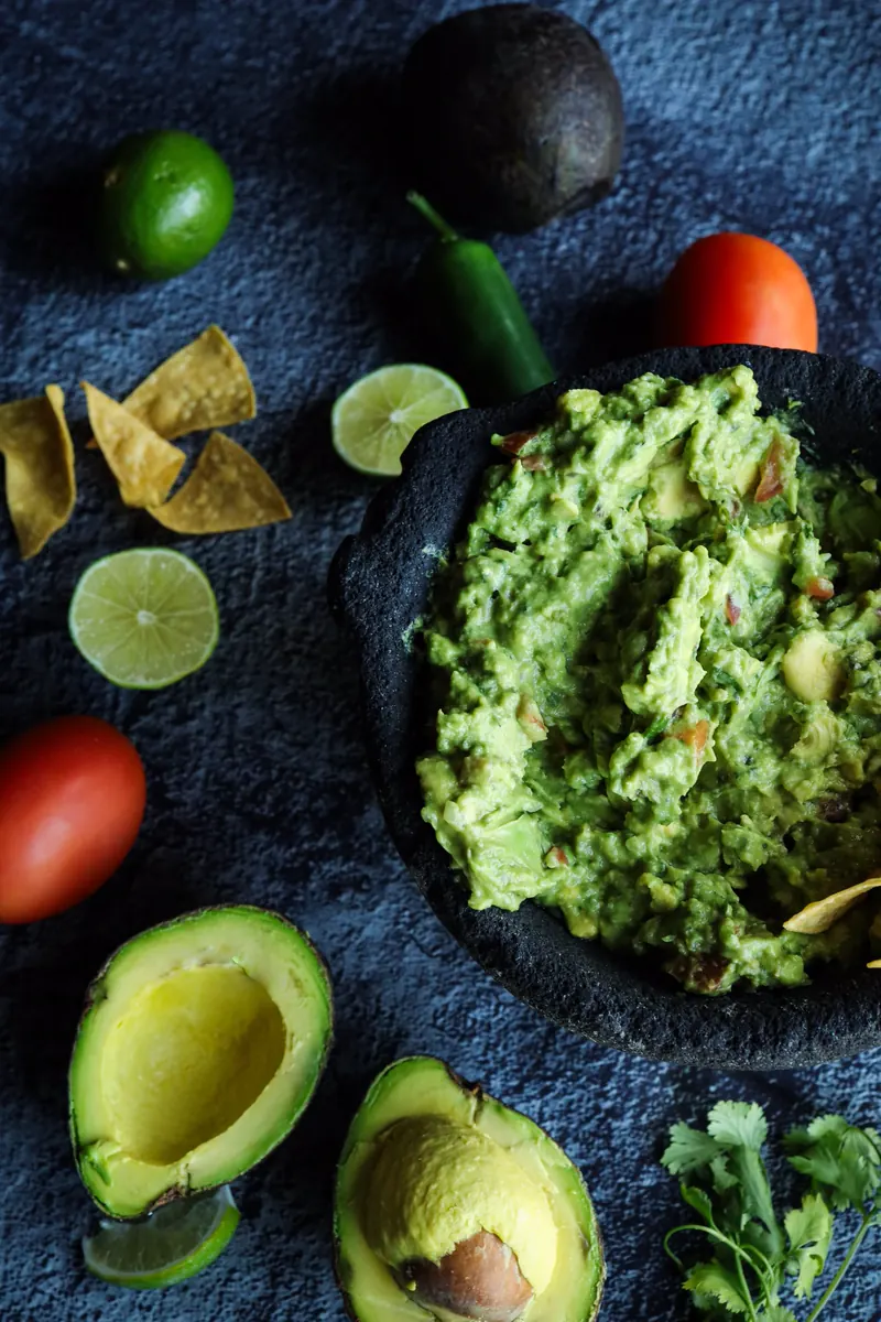 Guacamole in a black stone molcajete, surrounded by halved avocados, limes, tomatoes, a jalapeño and tortilla chips on blue-grey slate