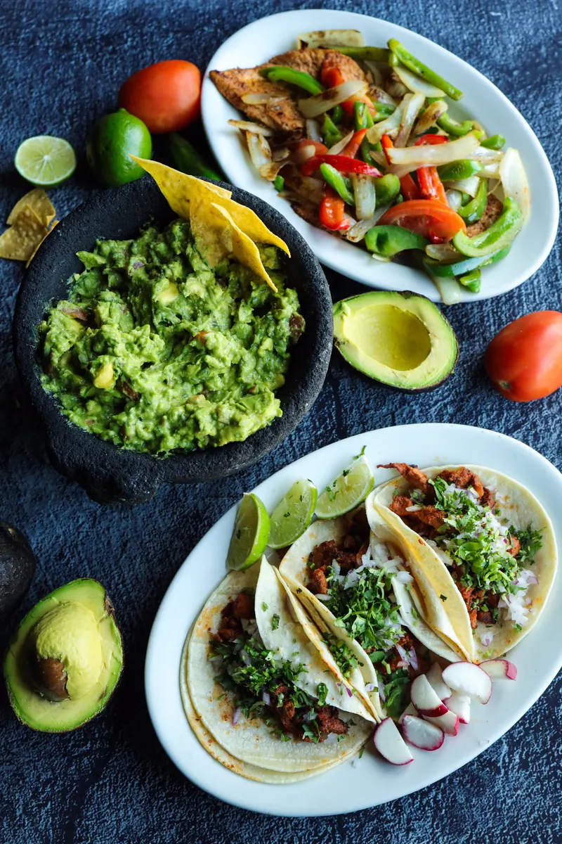 Flat-lay on slate: guacamole in the molcajete, a plate of sizzling fajitas and three tacos with radish and lime