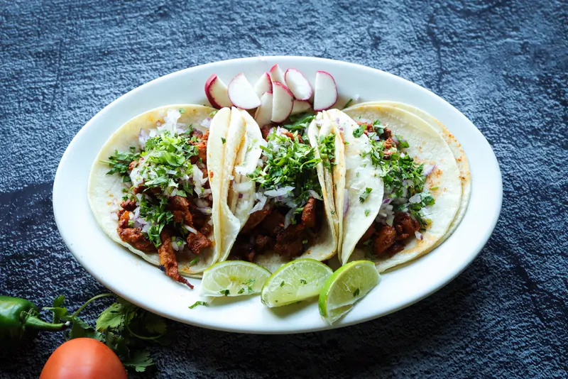 Three soft tacos with cilantro, onion and radish, with lime wedges on a white oval plate on slate