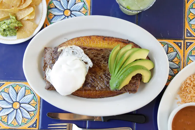 Bistec a la Criolla: grilled steak on a fried sweet plantain with an egg over easy and sliced avocado, on blue Talavera tile