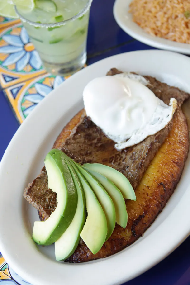 Bistec a la Criolla from above: steak on a fried plantain with a fried egg and fanned avocado, a margarita behind