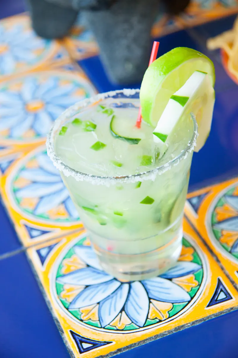 A green margarita with cucumber and jalapeño pieces, a salted rim and a lime wedge, on blue tile