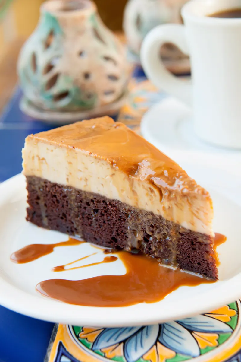 A slice of El Imposible, half chocolate cake and half flan, with caramel on a white plate and a cup of coffee behind