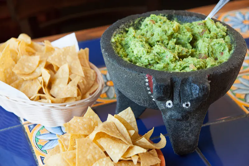 Guacamole in a stone molcajete carved like a bull, with a basket of tortilla chips on blue Talavera tile