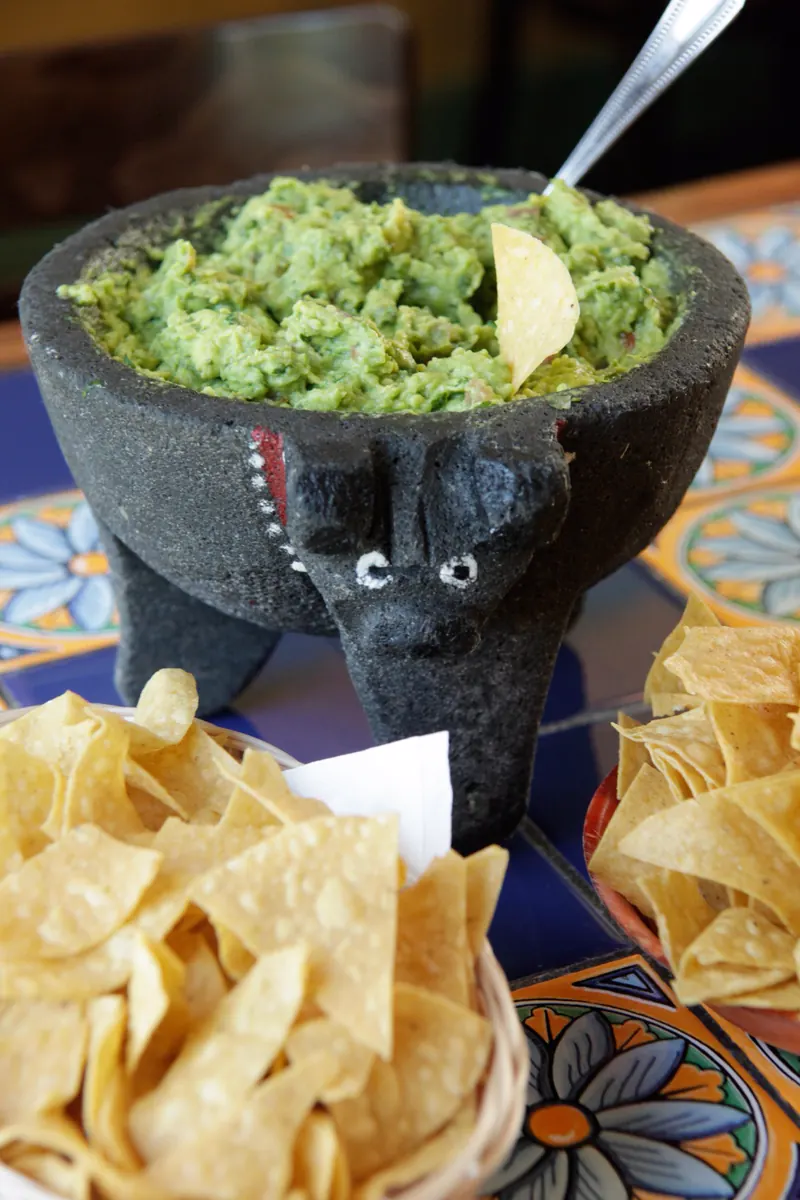 Guacamole in the stone molcajete with a basket of tortilla chips on blue tile