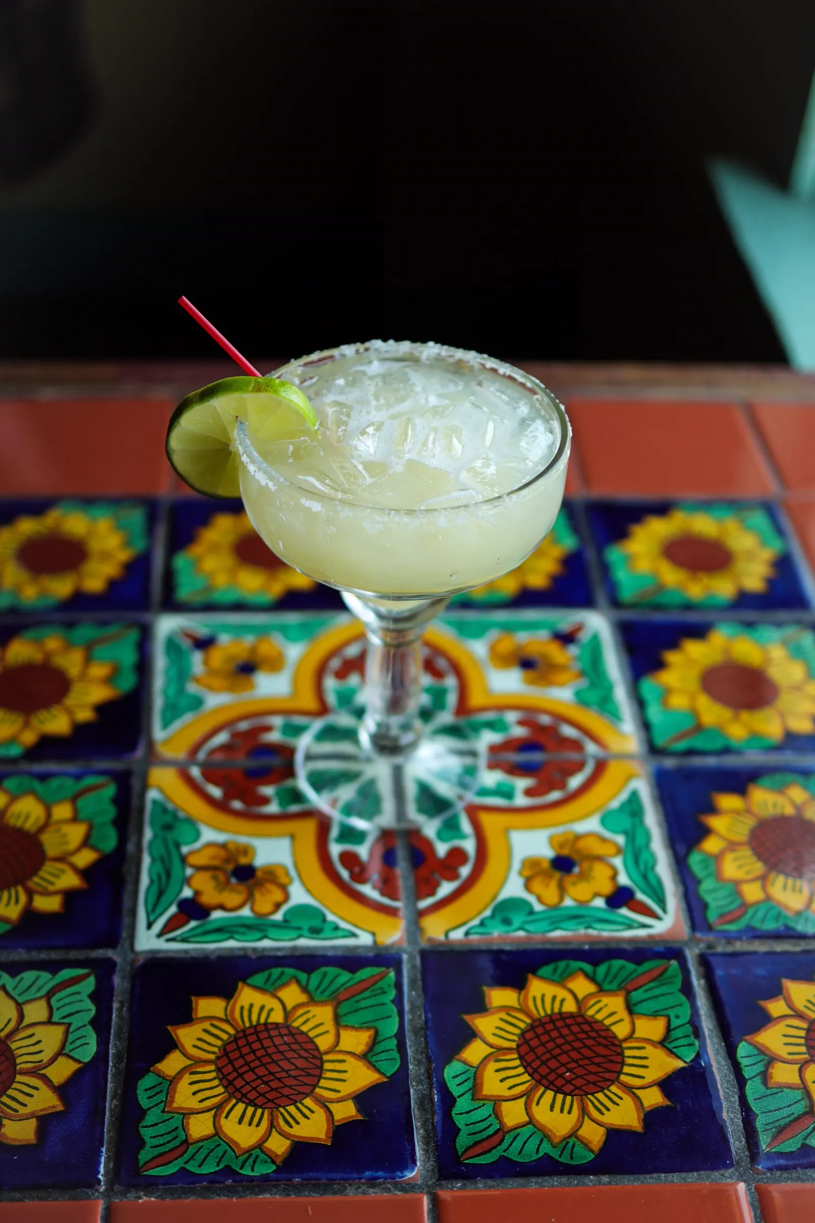 A fresh margarita with a salted rim and a lime wheel on a sunflower Talavera tile table, against a dark room