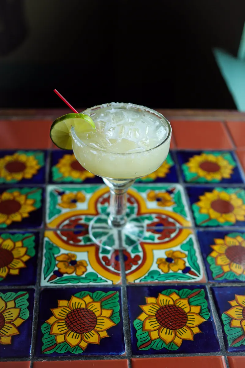 A margarita with a salted rim and a lime wheel on a table of sunflower-pattern Talavera tiles, against a dark room