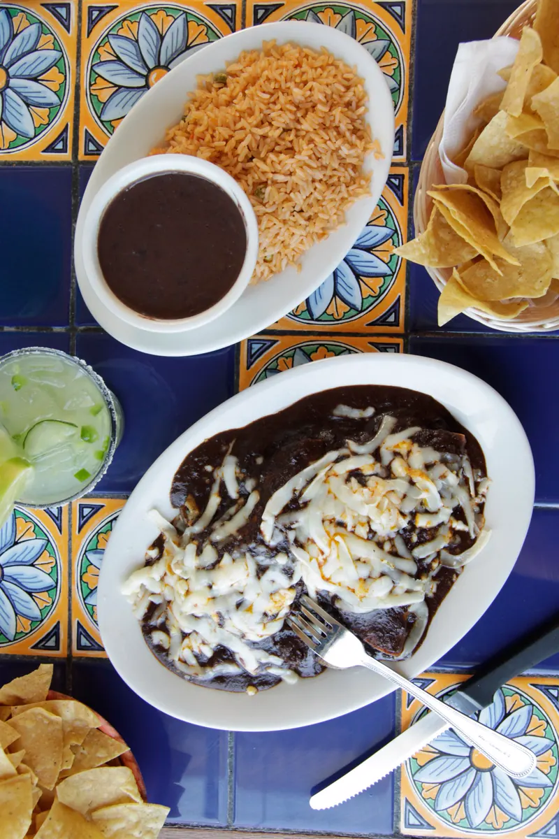 Enchiladas de mole poblano topped with melted cheese, with rice, black beans and a basket of chips on blue Talavera tile