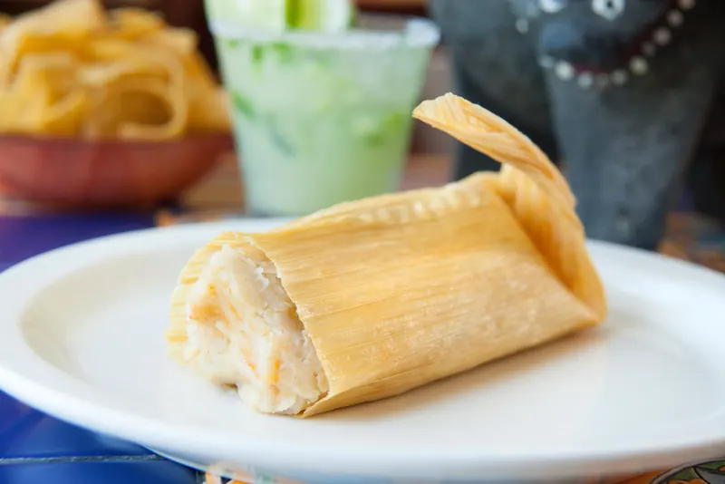 A homemade tamal in its corn husk on a white plate, with a margarita and the stone molcajete behind