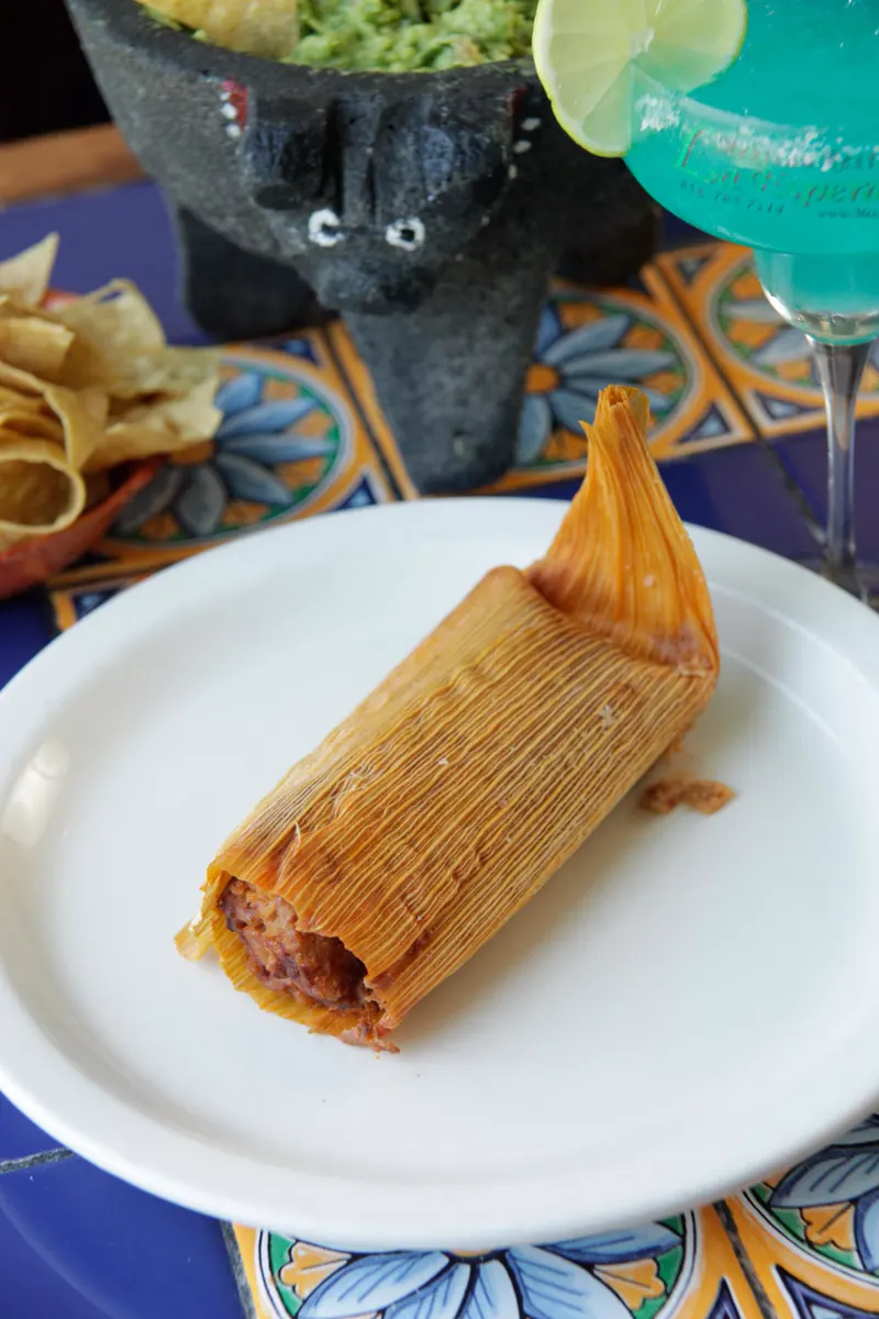 A tamal in its corn husk on a white plate with a blue margarita behind it