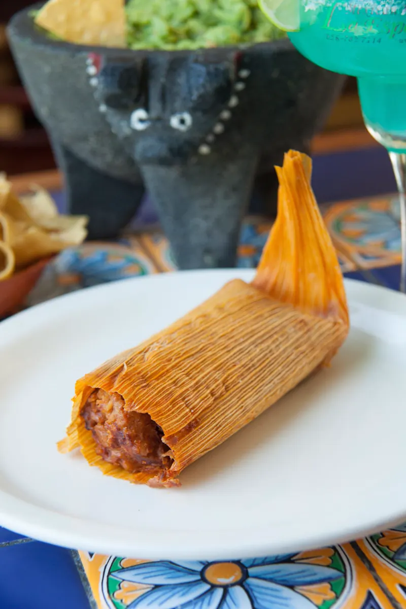 A homemade tamal opened in its corn husk on a white plate, the molcajete behind